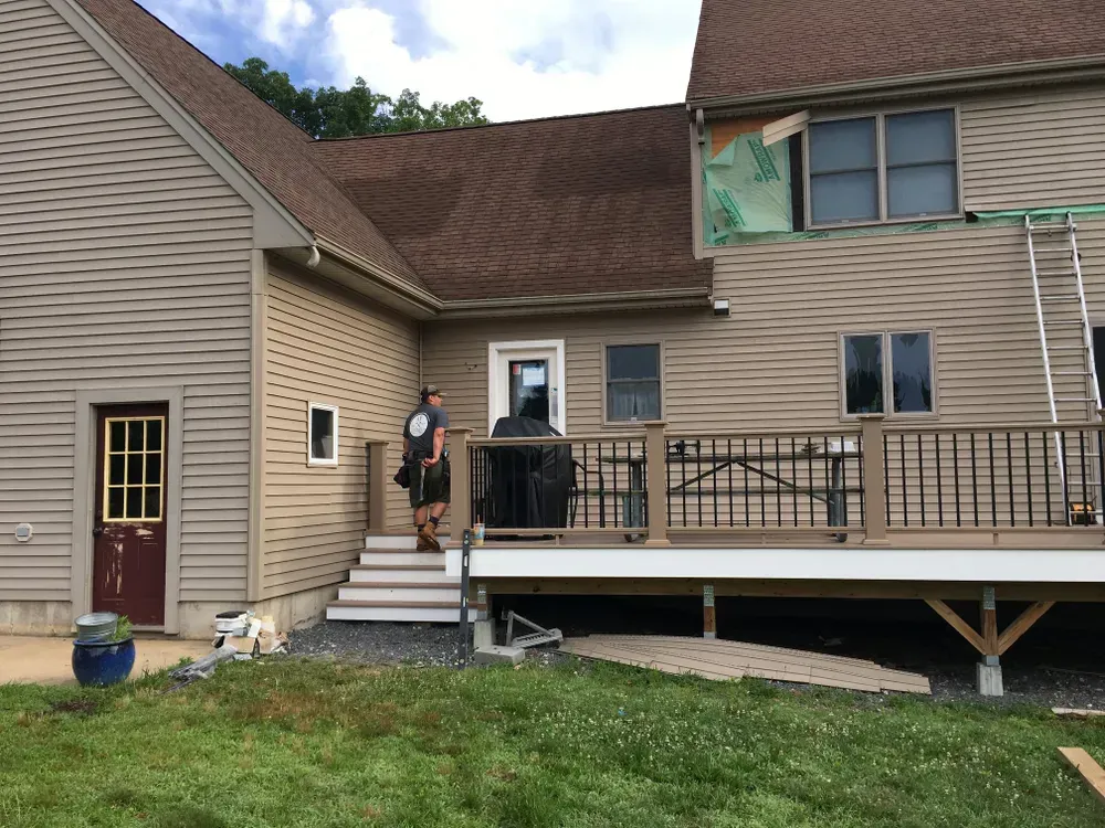 A person stands on the deck steps of a tan house with brown trim and a dark shingled roof under a cloudy sky.