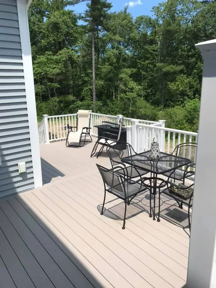 A light-colored outdoor deck with a glass dining table, four chairs, a lounge chair, and a grill, overlooking lush trees.