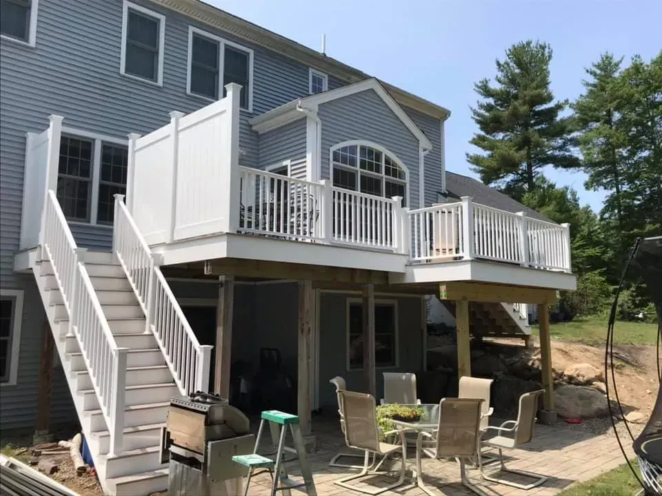 A multi-level light gray house with a large white deck, outdoor staircase, and a patio set underneath in the backyard.