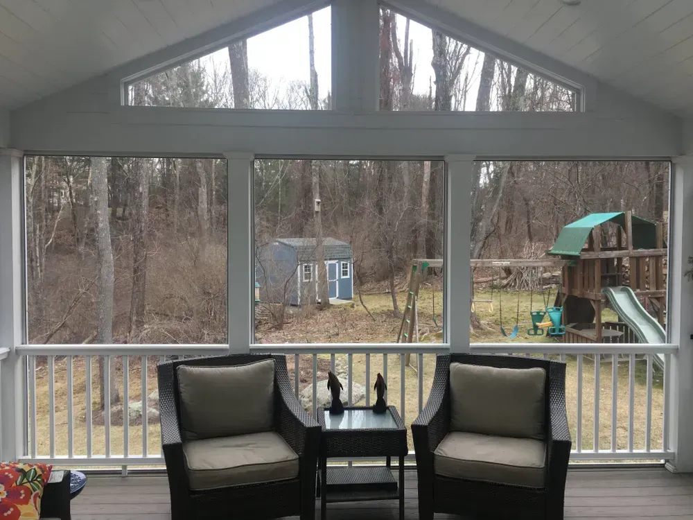 Two dark wicker chairs and a small table on a screened-in deck, overlooking a backyard with a shed and play set.