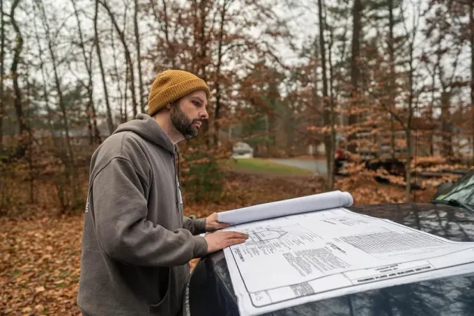 A person in a gray hoodie and yellow beanie reviews construction blueprints spread across the hood of a dark vehicle.