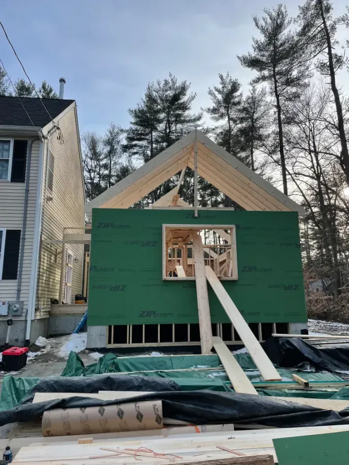 A house addition under construction with green exterior sheathing and a wooden gable roof frame, set near tall trees.