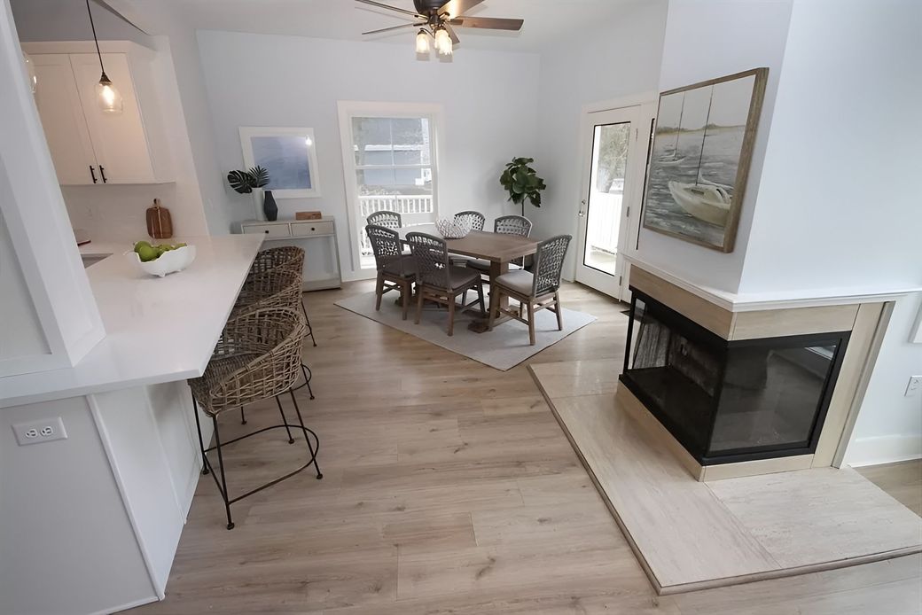 A bright, open-plan dining and kitchen area with a wooden table, woven chairs, a white breakfast bar, and a fireplace.