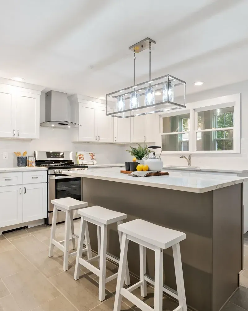 Modern kitchen featuring white cabinets, a grey island with three white stools, and a glass-paneled light fixture above.