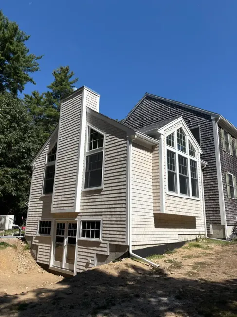 A light-colored, multi-story house addition with large windows and a chimney, situated next to an older dark-shingled home.