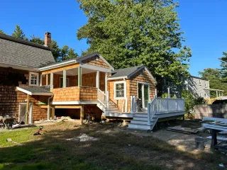 A house under construction with new wood-shingled additions, an open porch, and a white-railed deck on a sunny day.