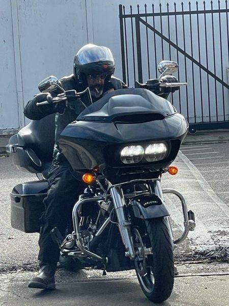 Person wearing helmet on black Harley-Davidson motorcycle with bright headlights, parked near a fence.