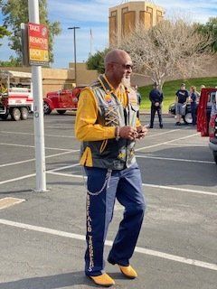 Man in yellow shirt and vest, blue jeans with side text, standing in parking lot, holding phone.