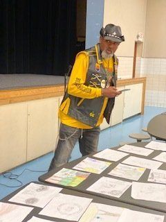 Person in yellow shirt and vest standing over a table of drawings. Stage background.