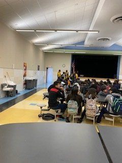 School cafeteria: Students seated at tables; stage at the back.