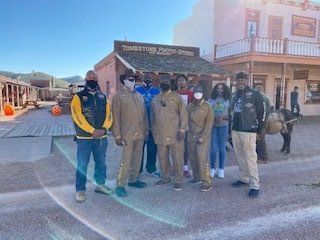 Group of people in front of a western-style building. Some wear masks, outfits with patches, and others casual clothes. Sunny day.