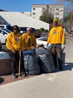 Three people in yellow hoodies stand with trash bags on a sidewalk, likely doing a cleanup.
