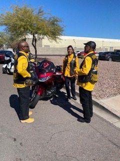 Three people in yellow vests stand near a red motorcycle in a parking lot on a sunny day.
