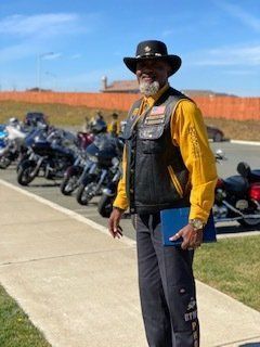 Man in biker vest and hat stands in front of parked motorcycles, holding a blue book, outdoors.