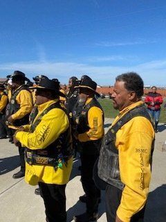 Men in yellow jackets and black vests stand outside, blue sky, some wearing hats.