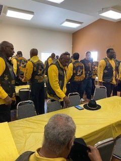 Men in yellow vests, standing in a room. Table with yellow cloth, black hat.