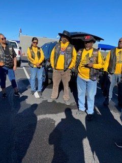 Group of people in vests and yellow shirts standing in parking lot, car behind.