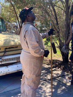 Person in light-colored jumpsuit looks up while holding a pole. Standing outside near a vehicle and trees.