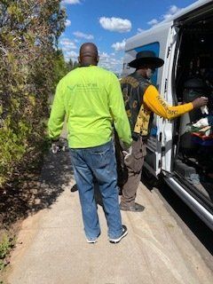 Two people near a van; one in green shirt and jeans, other in yellow and black, loading items.