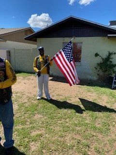 Man holding American flag in front of house. Sunny day, green grass, another person partially visible.