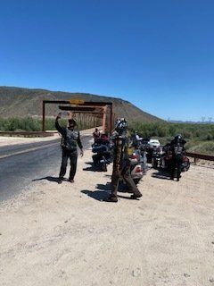 Motorcyclists by a bridge; two wave near parked bikes on a sunny day.