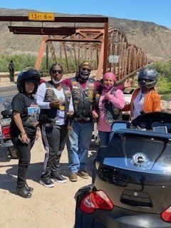 Group of people on motorcycles pose in front of a bridge. They wear vests, helmets, and smiles.