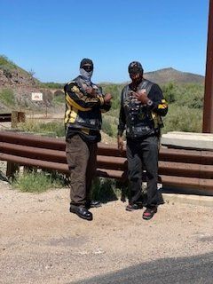 Two men in vests, standing outside with a brown fence and shrubs in the background. One wears a face covering.