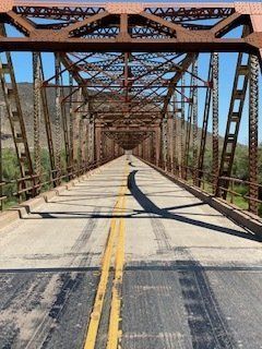 Rusty metal bridge with a paved road, mountains in the background, bright sunny day.