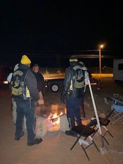 Group of people in jackets around a bonfire at night. Some have patches. Chairs nearby.