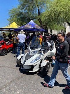 A white and black three-wheeled motorcycle parked outdoors with people and tents nearby on a sunny day.