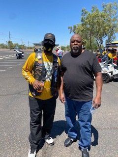 Two men posing outdoors near motorcycles; one wearing a yellow shirt and vest, the other in a black shirt and jeans.
