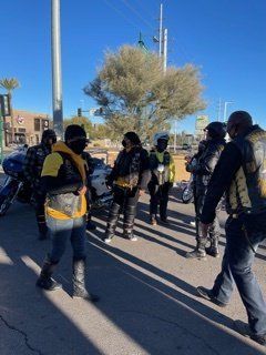 Group of people in black vests and helmets standing near parked motorcycles on a sunny street.