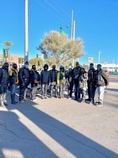 Group of people standing outside; sunny day; some wearing masks and jackets.