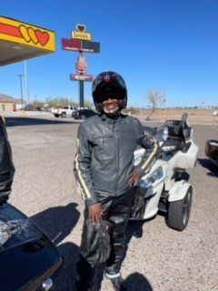 Person in motorcycle gear stands next to a three-wheeled vehicle outside a gas station on a sunny day.