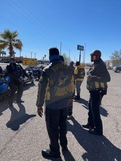 Group of people in jackets with a motorcycle club logo standing near motorcycles outside on a sunny day.