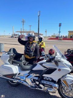 Two motorcyclists pose beside a white Honda Goldwing. One waves. Sunny outdoors, roadside setting.