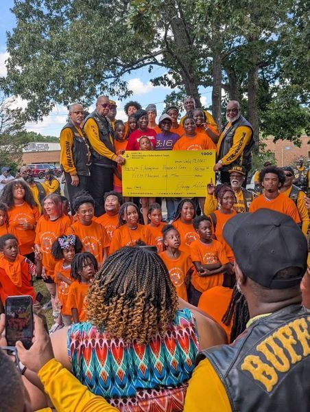 People presenting a large check to a group of children in orange shirts, outdoors.
