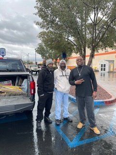 Three men stand next to a truck in a parking lot on a cloudy day. One man gestures with a peace sign.