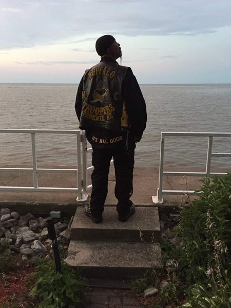 Person wearing a biker jacket, standing on steps, gazing at a body of water under a cloudy sky.