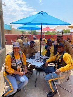 Group of people in matching jackets and hats sit around a table outdoors under blue umbrellas.