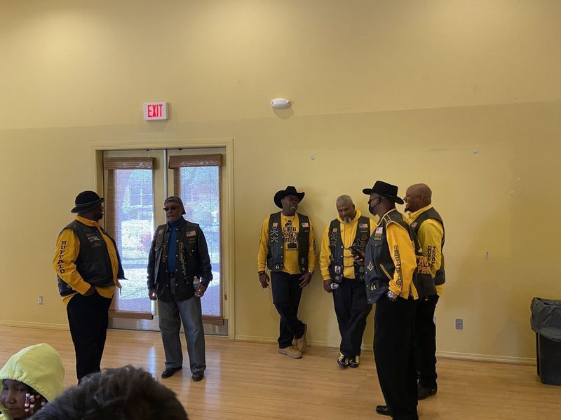 Group of men in vests and hats stand in a room with a wooden floor, near a door.