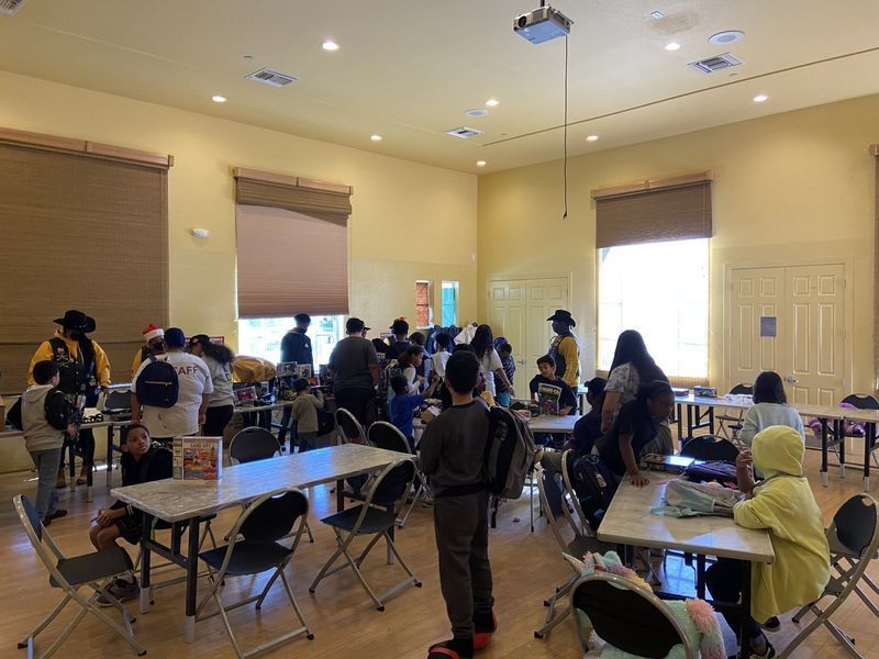 People gather in a room with tables and chairs, under tan-colored walls and window blinds.