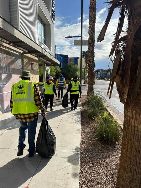 People in yellow vests pick up trash on a sidewalk, near a building and a street with palm trees.