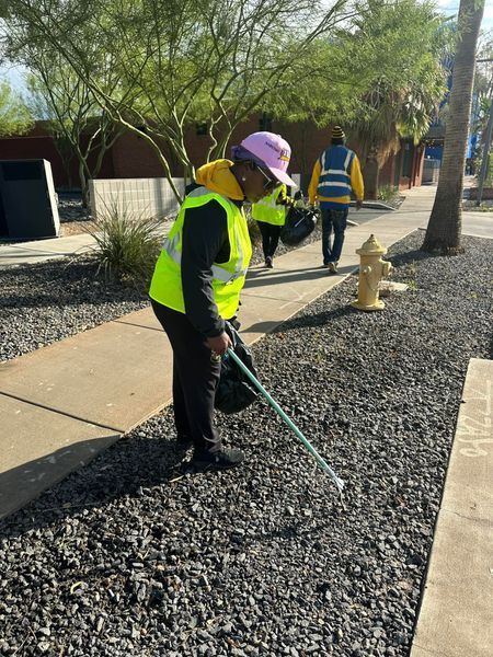 Person in yellow vest picks up trash along a sidewalk, others in background.