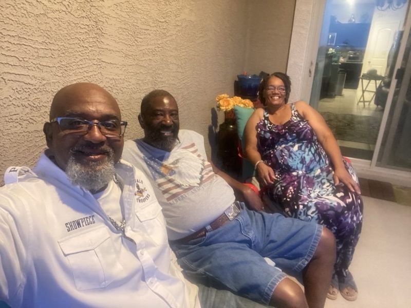 Three people seated on a couch outside. One man in white jacket, two others in casual attire, smiling.
