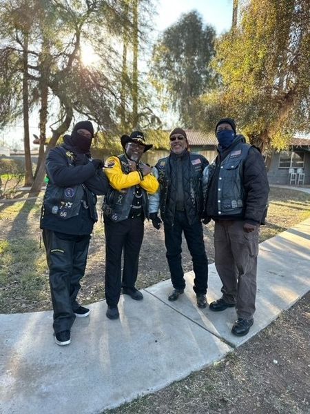 Four people in biker vests stand on a sidewalk, posing for a photo.