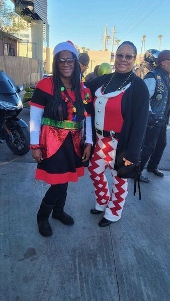 Two people in festive outfits pose outdoors. One wears elf attire, the other a harlequin style.
