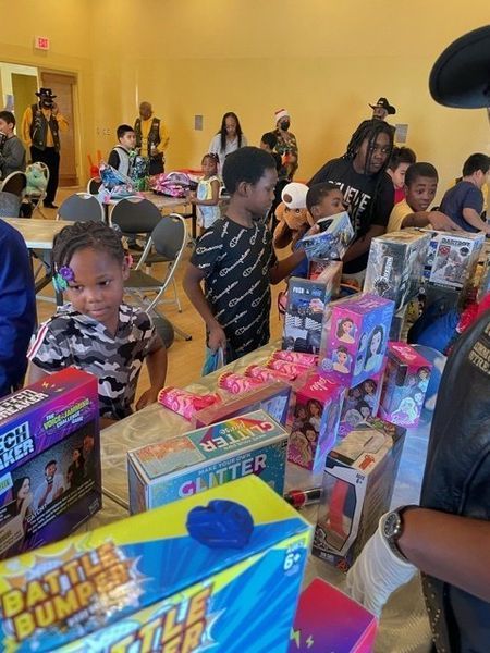 Children selecting toys from tables at an event. Boxes of toys in foreground, people in the background.