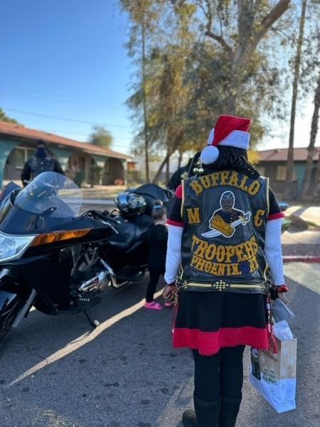 Person in Santa hat, Buffalo Troopers MC vest, stands by motorcycles, holding gifts in Phoenix.