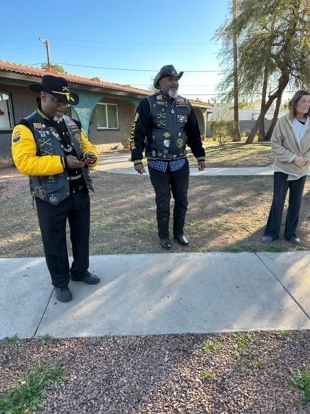 Two men in vests and a woman stand on a sidewalk near a house.